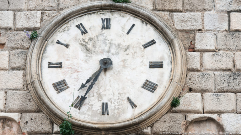 Large weathered white clock face with black Roman numerals and dark hands set into a pale stone wall.