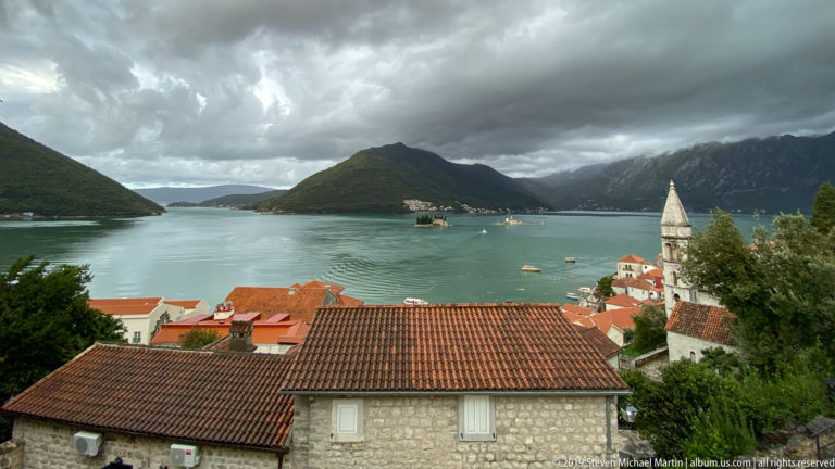 Elevated view over red-tiled Kotor roofs and a stone bell tower toward the winding Bay of Kotor and surrounding mountains.
