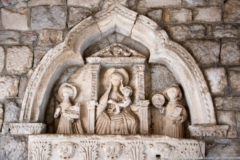 Stone relief beneath a pointed arch showing the seated Virgin and Child flanked by two standing figures.