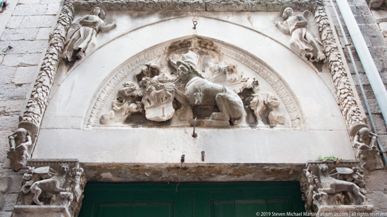 Ornate carved stone lunette with figures, heraldic motifs, and foliage above a green doorway in Kotor.