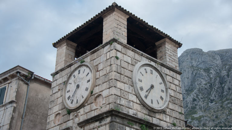 Stone clock tower with two large Roman-numeral faces and a dark roof, backed by steep limestone mountains in Kotor.