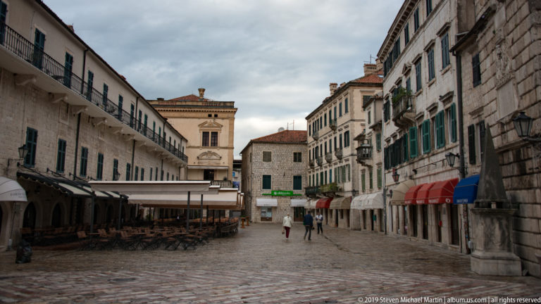 Pedestrian square in Kotor lined with pale stone buildings, striped awnings, balconies, arcades, and outdoor café tables.