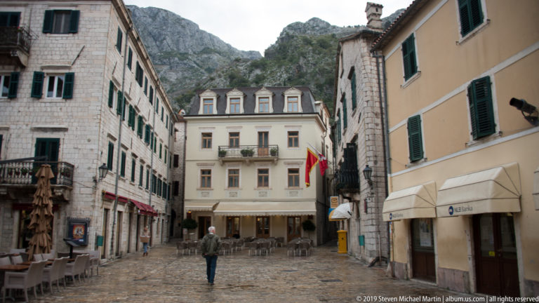 Cobbled pedestrian street lined with stone buildings, green shutters, café seating, and steep mountains rising behind Kotor Old Town.
