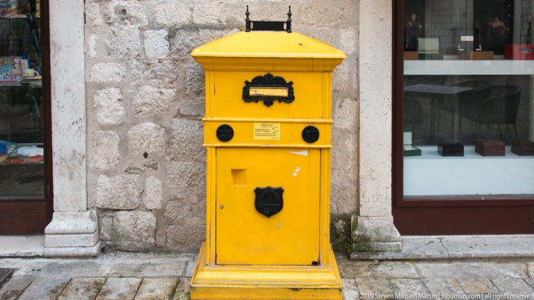 Bright yellow metal post box mounted on a pedestal against a pale stone wall beside a shop window in Kotor.
