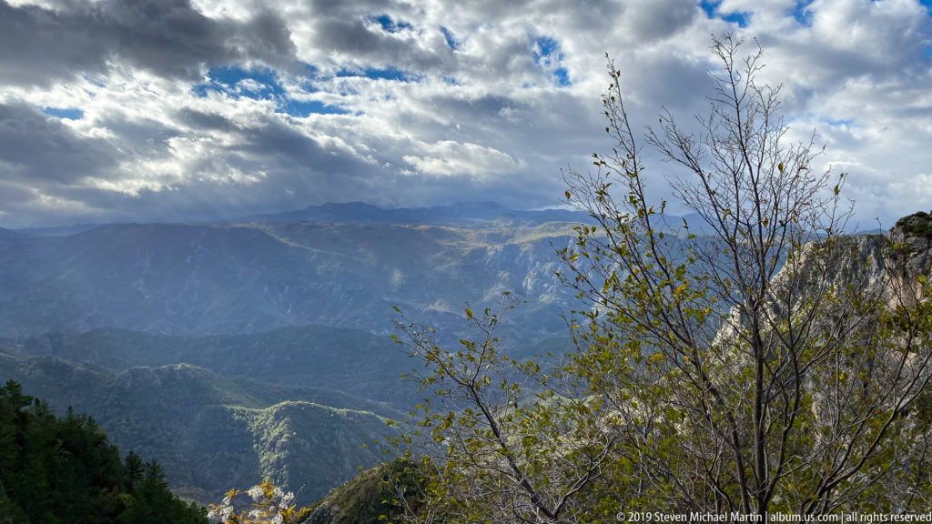 Layered gray limestone mountains under dramatic clouds, with sparse branches and sunlit ridges in the foreground and distance.