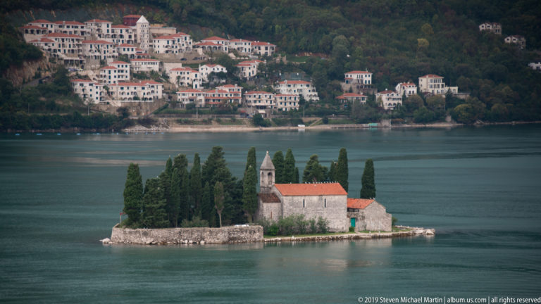 St. George Island with cypress trees and a stone church lies in front of hillside houses across the Bay of Kotor.
