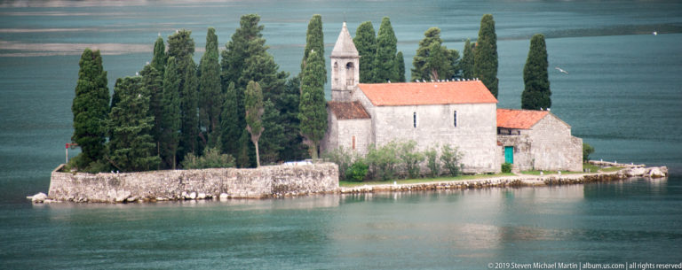 Stone church and monastery on St. George Island surrounded by tall cypress trees and enclosed by a low waterfront wall.
