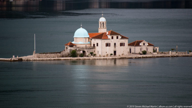 Close view of Our Lady of the Rocks, showing its blue dome, bell tower, pale walls, red roofs, and stone quay.