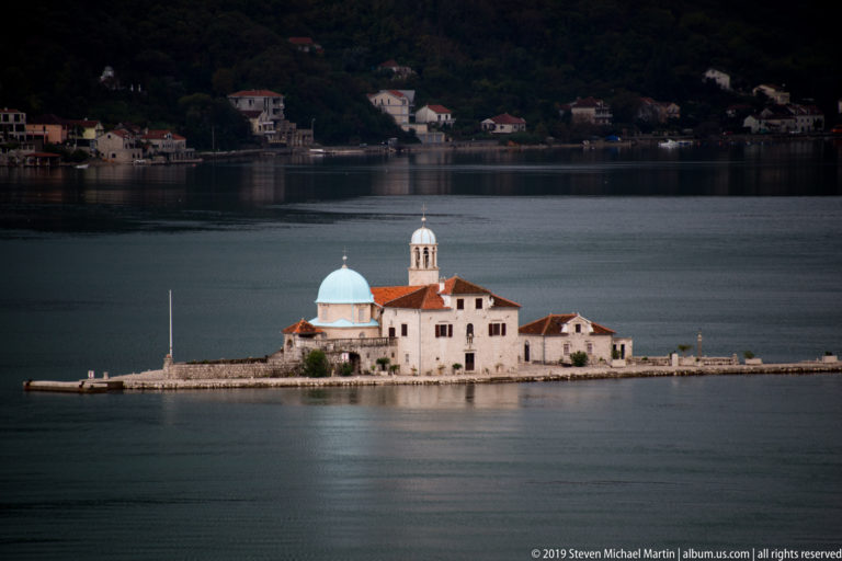 Our Lady of the Rocks church on a low stone island, with a blue dome, bell tower, red roofs, and dark bay behind.