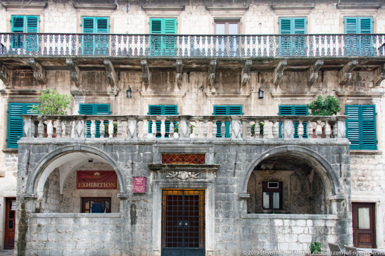 Historic stone palace façade with arcaded ground floor, balustraded balconies, green shutters, and an exhibition sign.