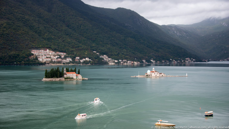 Two small church islands lie in the Bay of Kotor below forested mountains, with boats crossing calm green-blue water.