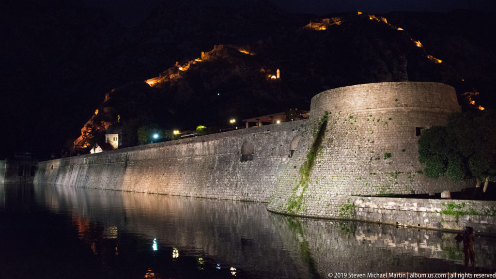 Long stone city wall and rounded bastion beside dark water, with illuminated Kotor fortifications climbing the mountain behind.