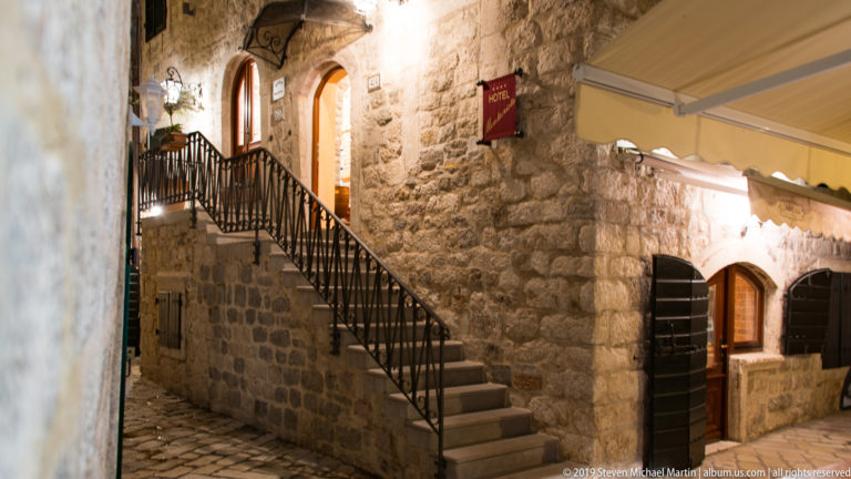 Stone stairway rising beside a rough masonry wall and restaurant awning on a narrow Kotor lane at night.