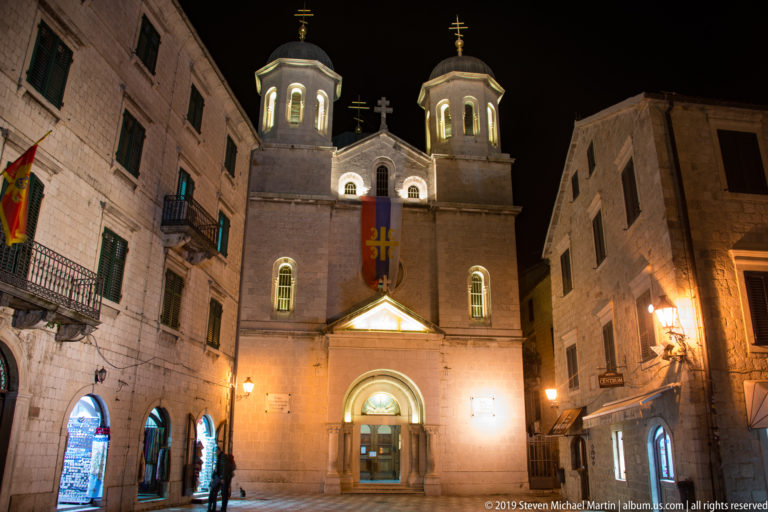 Full night façade of St. Nicholas Church in Kotor with twin domed towers, Orthodox crosses, and surrounding Old Town buildings.