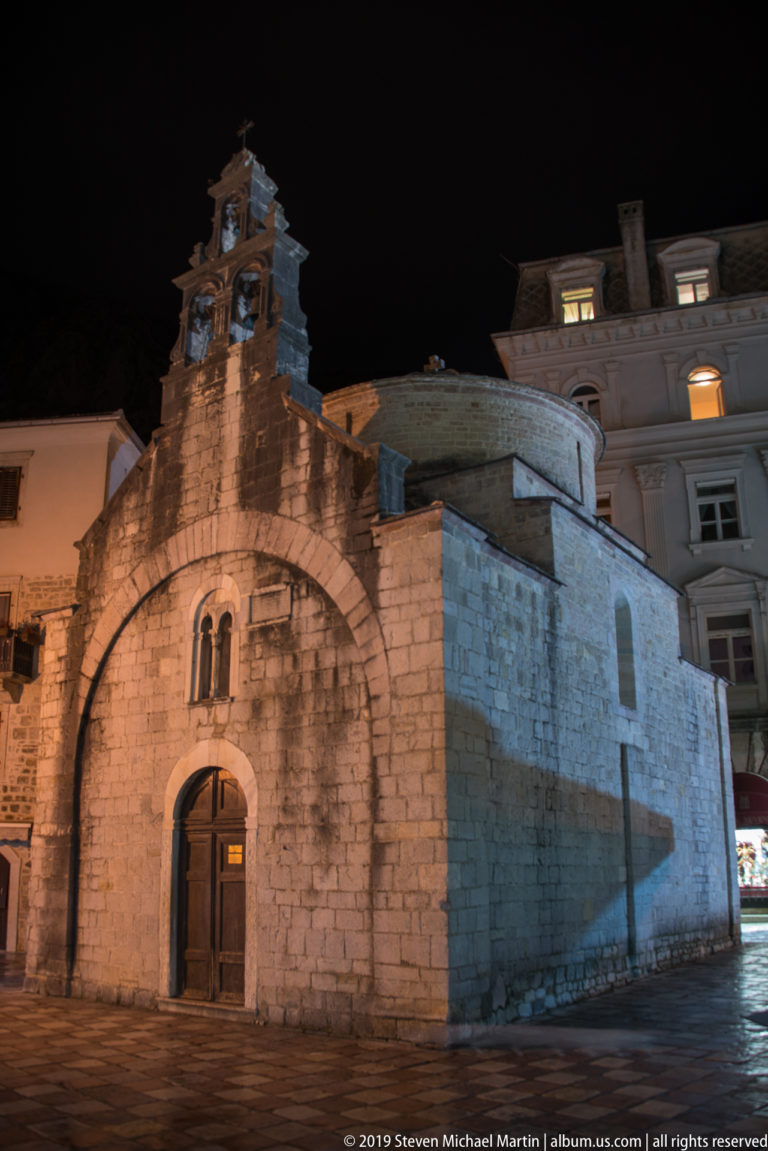 Oblique night view of the Church of St. Luke showing its bell gable, entrance façade, and plain stone side wall.