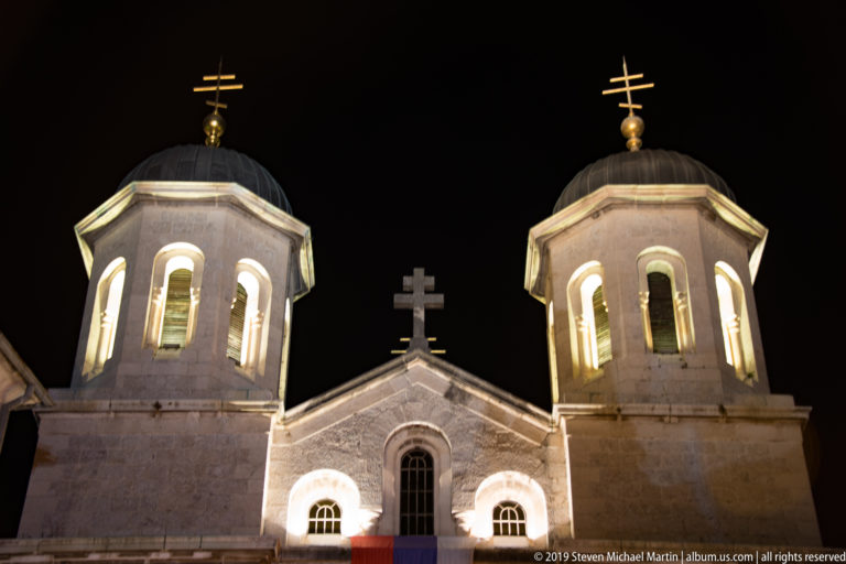 Illuminated twin domed bell towers and Orthodox crosses of St. Nicholas Church against a black night sky.