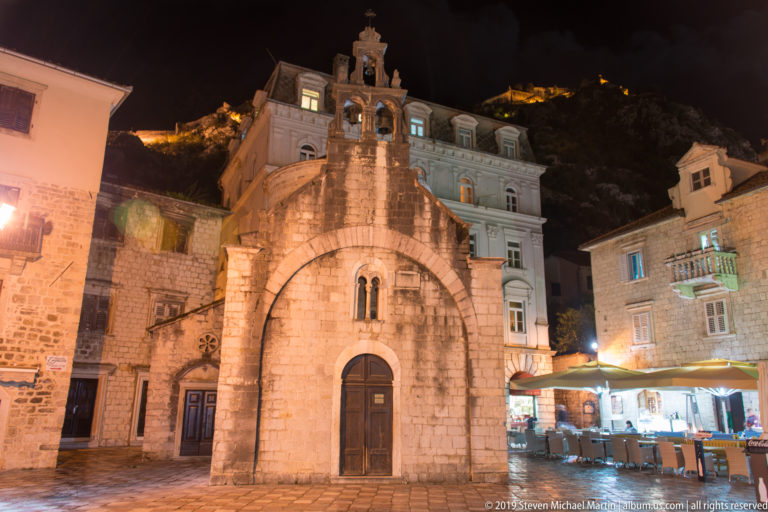 Church of St. Luke in Kotor illuminated at night, with a rounded doorway, small paired window, and bell gable.
