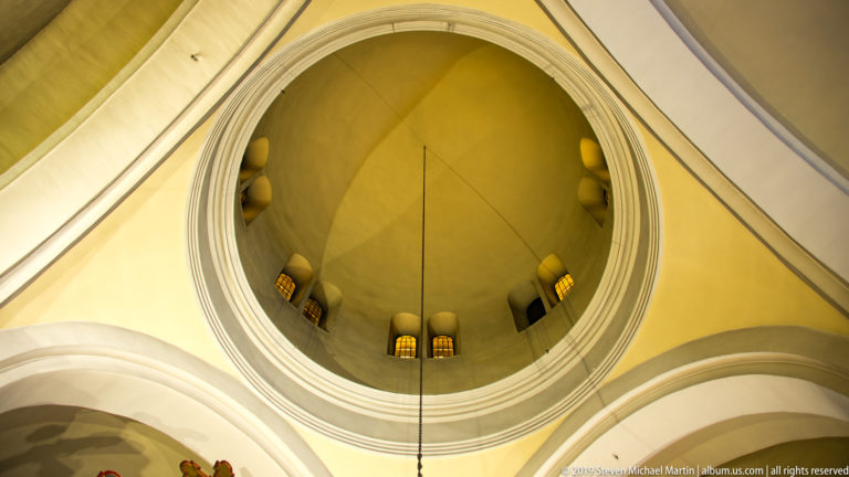 Upward view into the yellow central dome of St. Nicholas Church with small windows around the drum and white arches below.