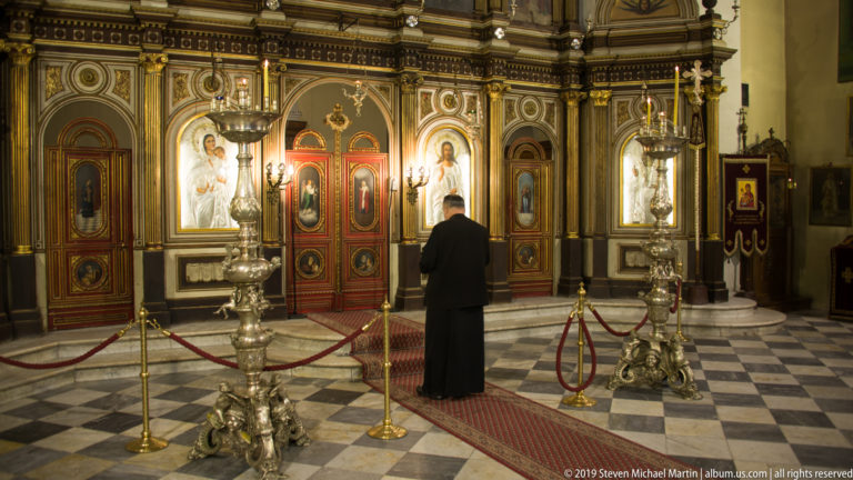 Person standing before the ornate iconostasis of St. Nicholas Church, with tall candle stands on a checkered floor.