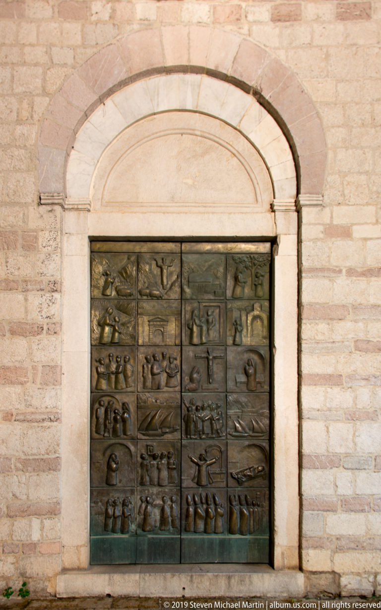 Bronze double doors covered with rows of narrative relief panels set within a pale stone arched portal.