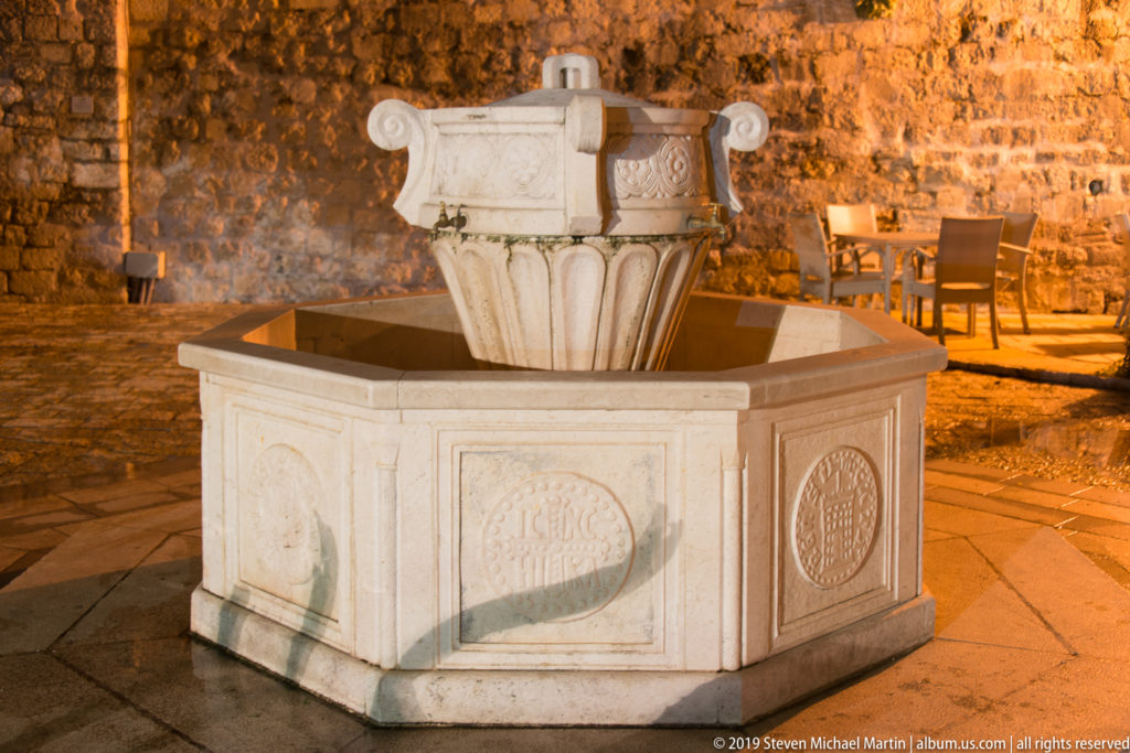 Octagonal pale-stone fountain with carved roundels and a fluted central pedestal, illuminated at night in Kotor.
