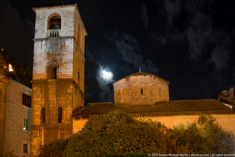 Square stone church tower and rounded apse illuminated at night beneath clouds and moonlight in Kotor.