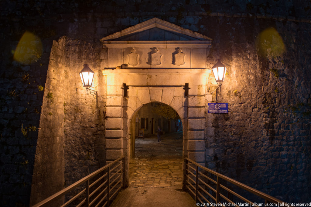 Illuminated stone gate with rusticated blocks, triangular pediment, and bridge railings in Kotor’s defensive wall.