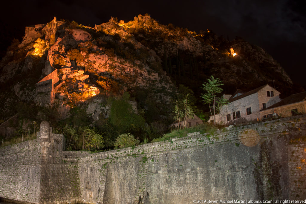 Illuminated sections of Kotor’s hillside defensive walls climbing a dark rocky slope above stone buildings.