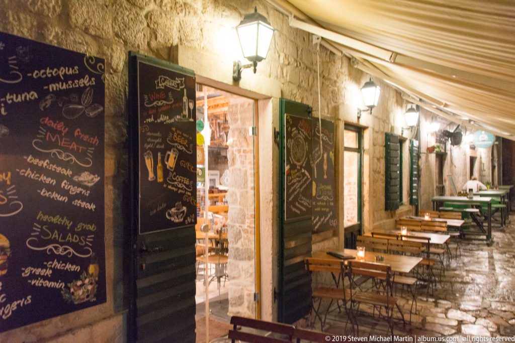 Outdoor café tables beneath an awning on a narrow stone lane with menu boards and wall lanterns in Kotor.