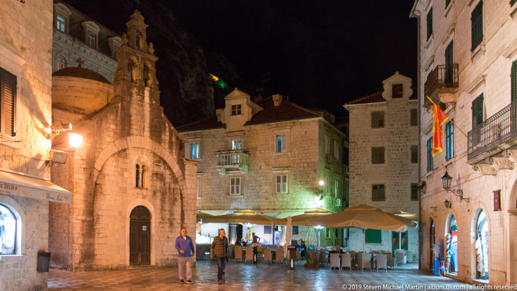 Small stone church beside cafés and pedestrians in a warmly lit Kotor Old Town square at night.