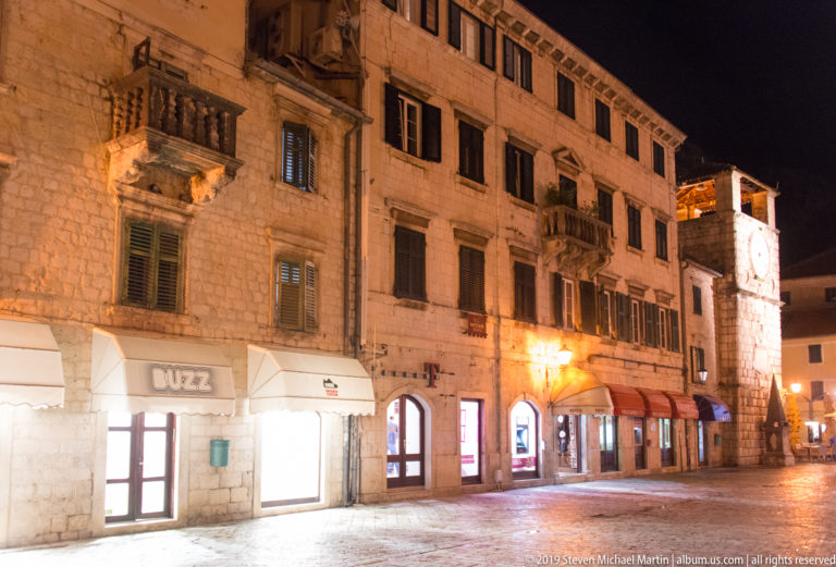 Night street in Kotor with illuminated shops, stone façades, balconies, shutters, and the Clock Tower at right.