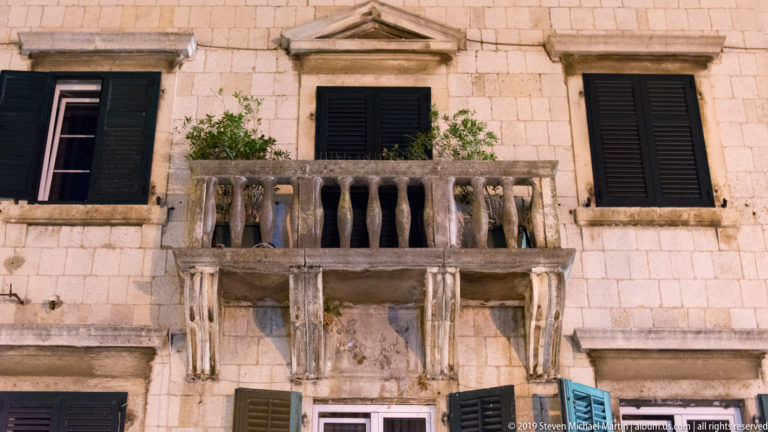 Stone balcony with balusters, black shutters, and classical window pediments on a historic Kotor façade.