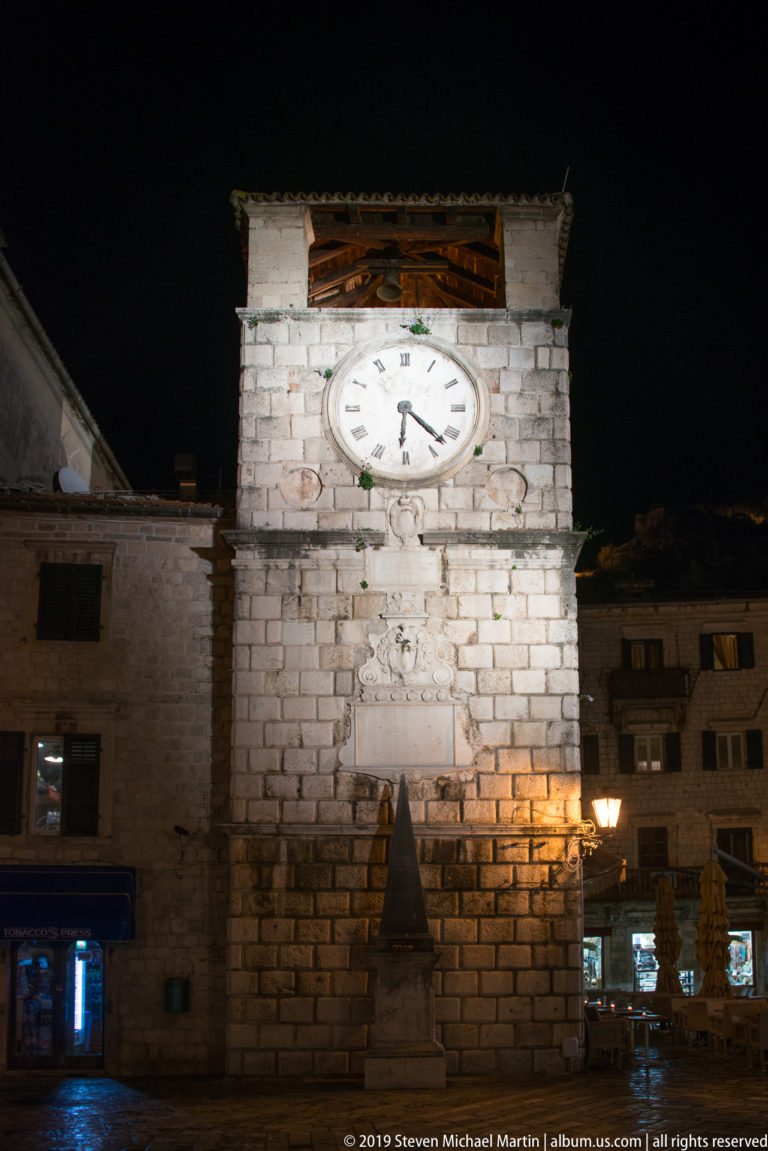 Frontal night view of Kotor Clock Tower with one bright clock face above the stone façade and lamps at street level.