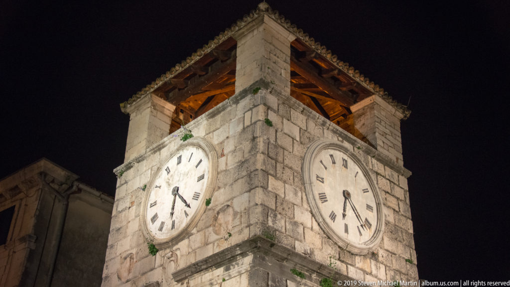 Low-angle night view of Kotor Clock Tower showing two illuminated clock faces and the open timber structure beneath its roof.