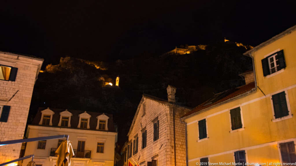 Illuminated stone houses in Kotor Old Town below scattered lights marking hillside fortifications on Mount St. John.