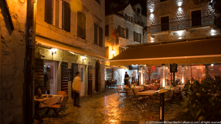 Outdoor restaurant tables beneath an awning on a warmly lit stone street in Kotor Old Town at night.