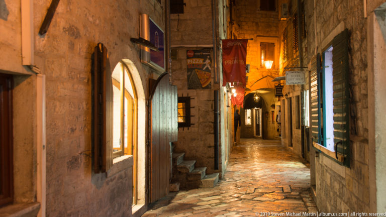 Narrow stone-paved lane with arched shopfronts, steps, signs, and warm lights in Kotor Old Town at night.