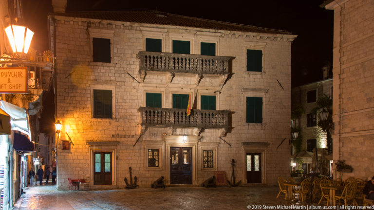 Three-story stone palazzo with two balustraded balconies, shuttered windows, and a central doorway illuminated at night in Kotor.