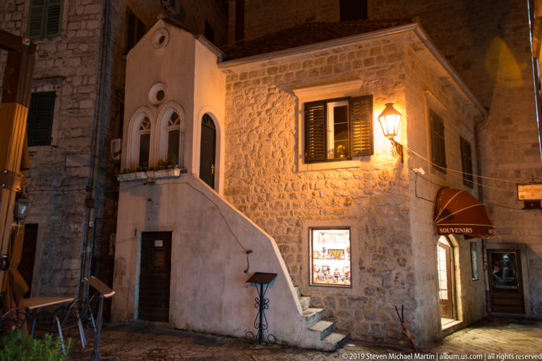 Stone house with shuttered windows, exterior stair, lantern, and small shop on a narrow Kotor Old Town street at night.