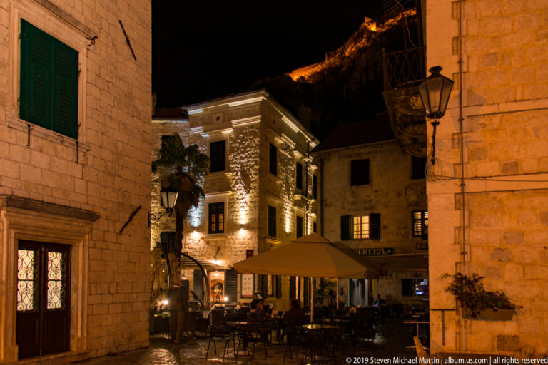 Outdoor café tables between illuminated stone buildings in Kotor, with hillside fortifications glowing above in the background.