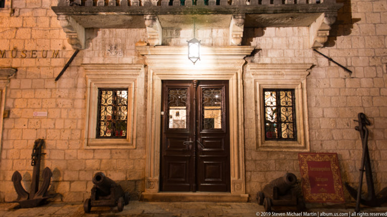 Night entrance of the Maritime Museum of Montenegro in Kotor, with carved stone portal, barred windows, anchors, and cannon.