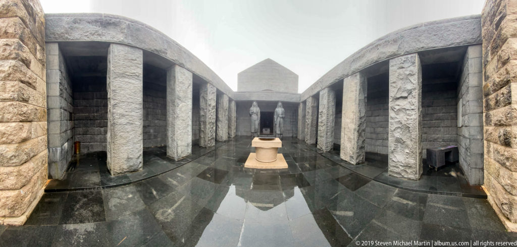 Wide-angle stone forecourt with wet reflective floor, central circular feature, and paired female guardian statues at the far end.