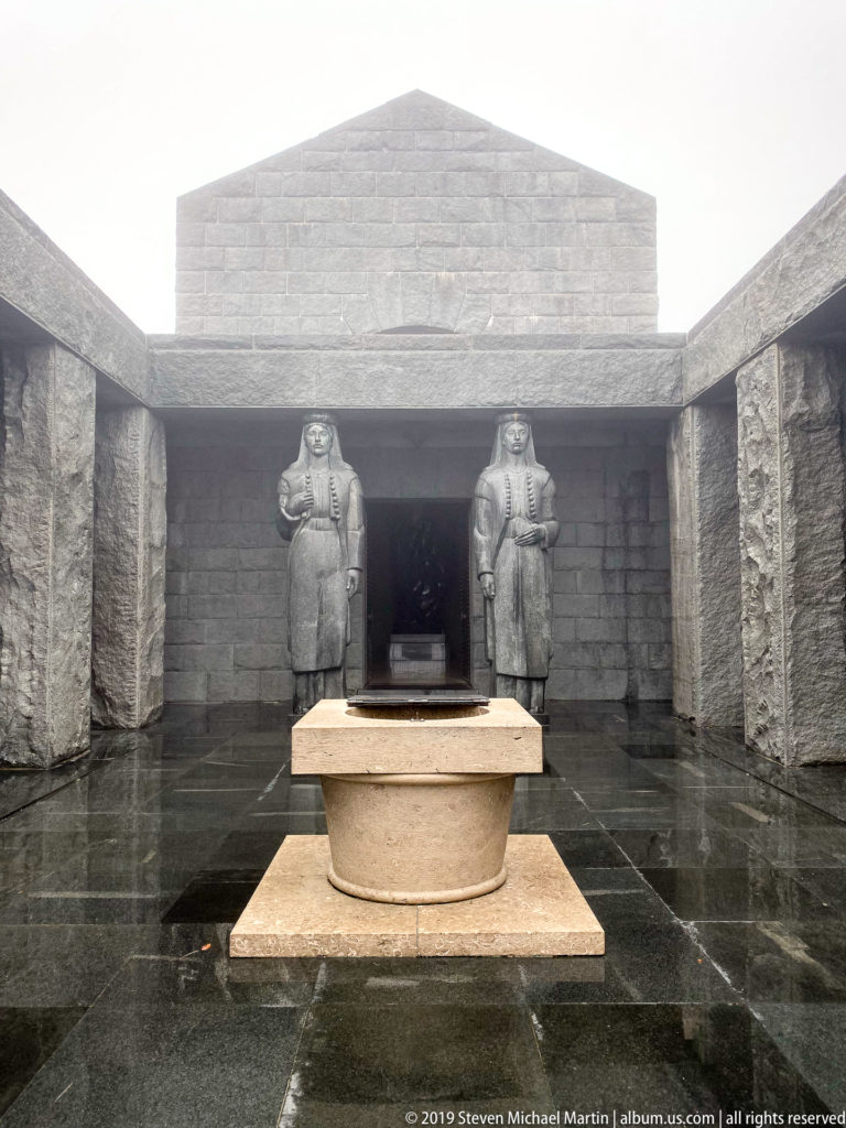 Two monumental gray stone women flank a dark mausoleum doorway beyond a low circular stone element on wet paving.
