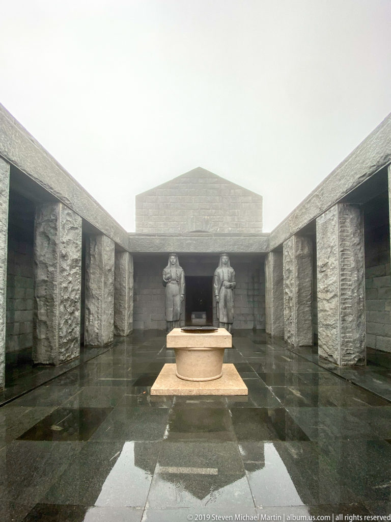 Low circular stone feature centered on wet paving before two monumental female statues at the mausoleum entrance.