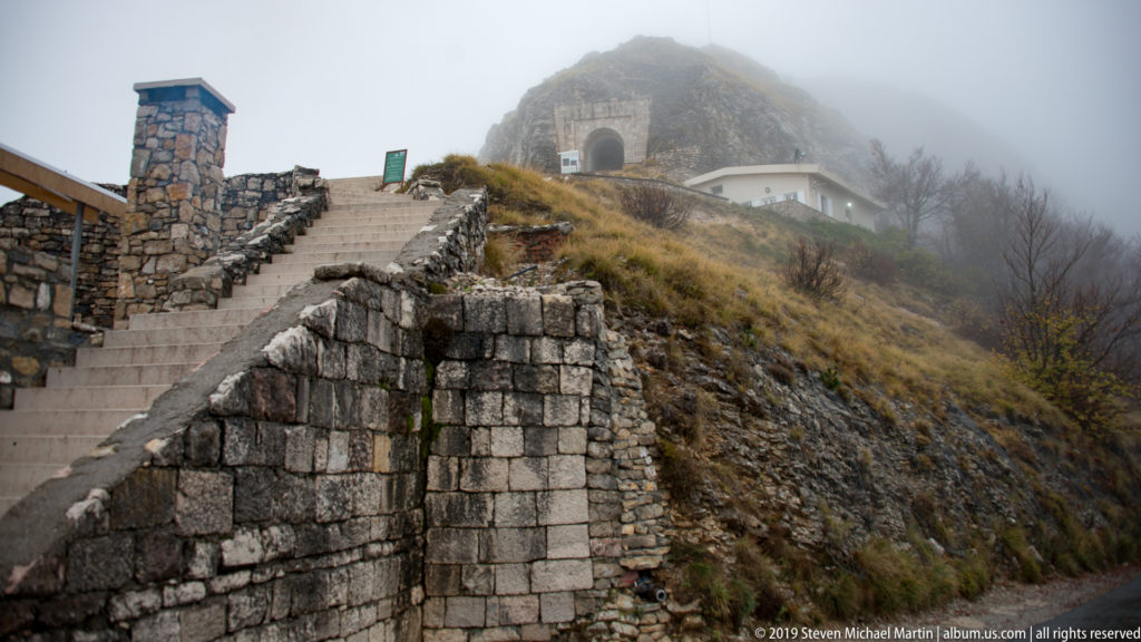 Stone staircase climbing beside masonry walls toward a fog-shrouded arched portal on the rocky Lovćen summit.
