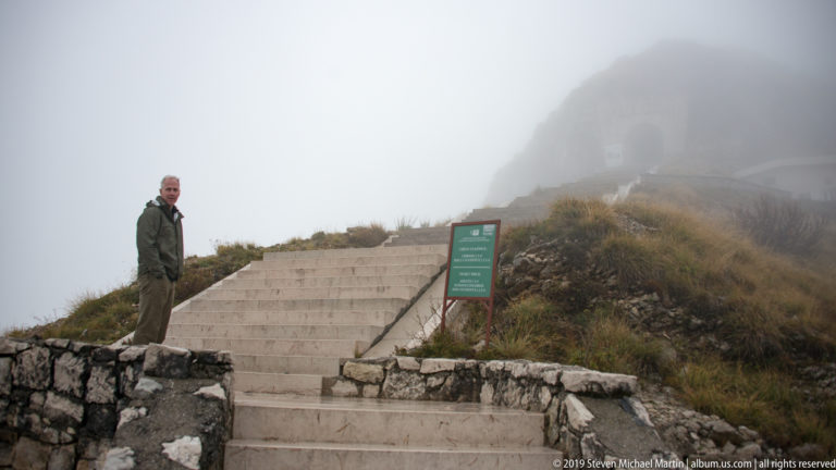 Man standing beside a green information board at the foot of stone steps that climb into dense fog toward the tunnel.