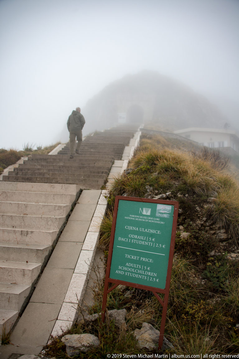 Green visitor information sign beside a broad stone staircase, with a person climbing into dense fog above.
