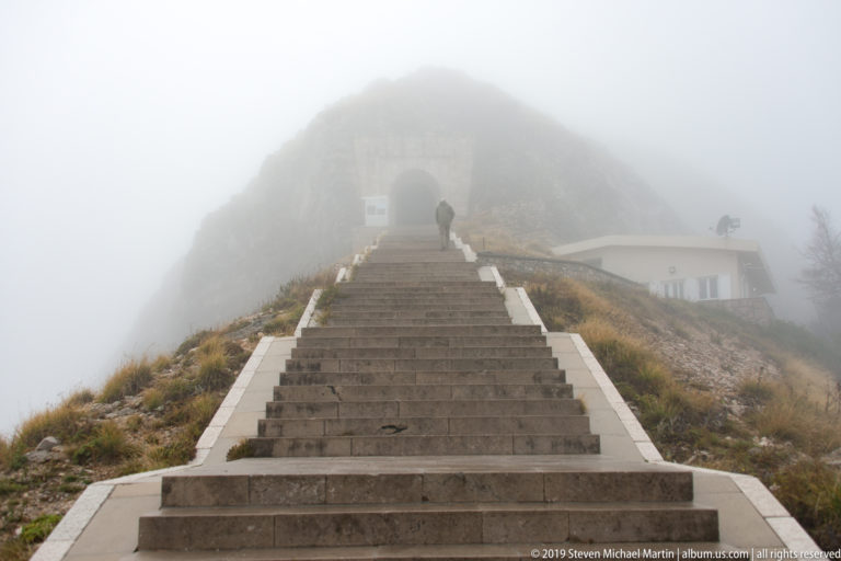 Long stone staircase disappearing into dense fog toward an arched tunnel entrance, with one visitor climbing near the top.