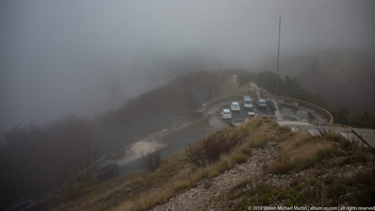 Foggy mountain arrival area with a circular road, parked vehicles, flagpole, dark trees, and tawny grass.
