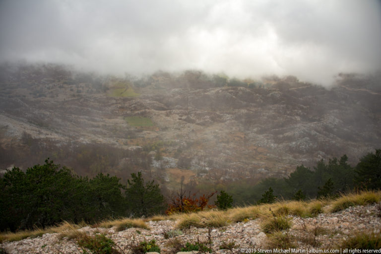 Low cloud drifting over a broad rocky limestone slope above dark conifers and tawny foreground grass.